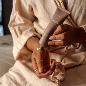 A person in a beige linen robe squeezes a tube of hand cream onto their palm. The tube is a muted taupe-brown colour with white text reading 'Barossa'