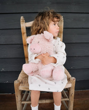 Child holding a pink plush soft toy pig sitting on a wooden chair against a dark wooden wall.