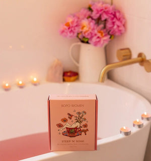 Box of 'Steep 'N' Soak' blushing bath tea on a bathtub with candles and flowers in the background