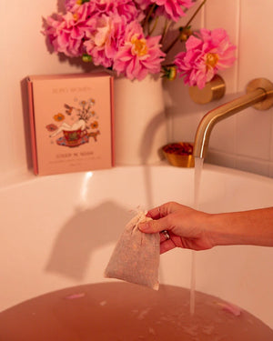 Person holding steep 'n' soak blushing bath tea bag over a bathtub with pink flowers and soap in the background