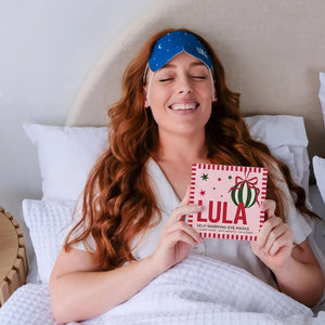 A smiling woman with long red hair lies in bed holding a box of self-warming eye masks. She is wearing a blue sleep mask and a white robe, with white pillows and a beige headboard behind her.