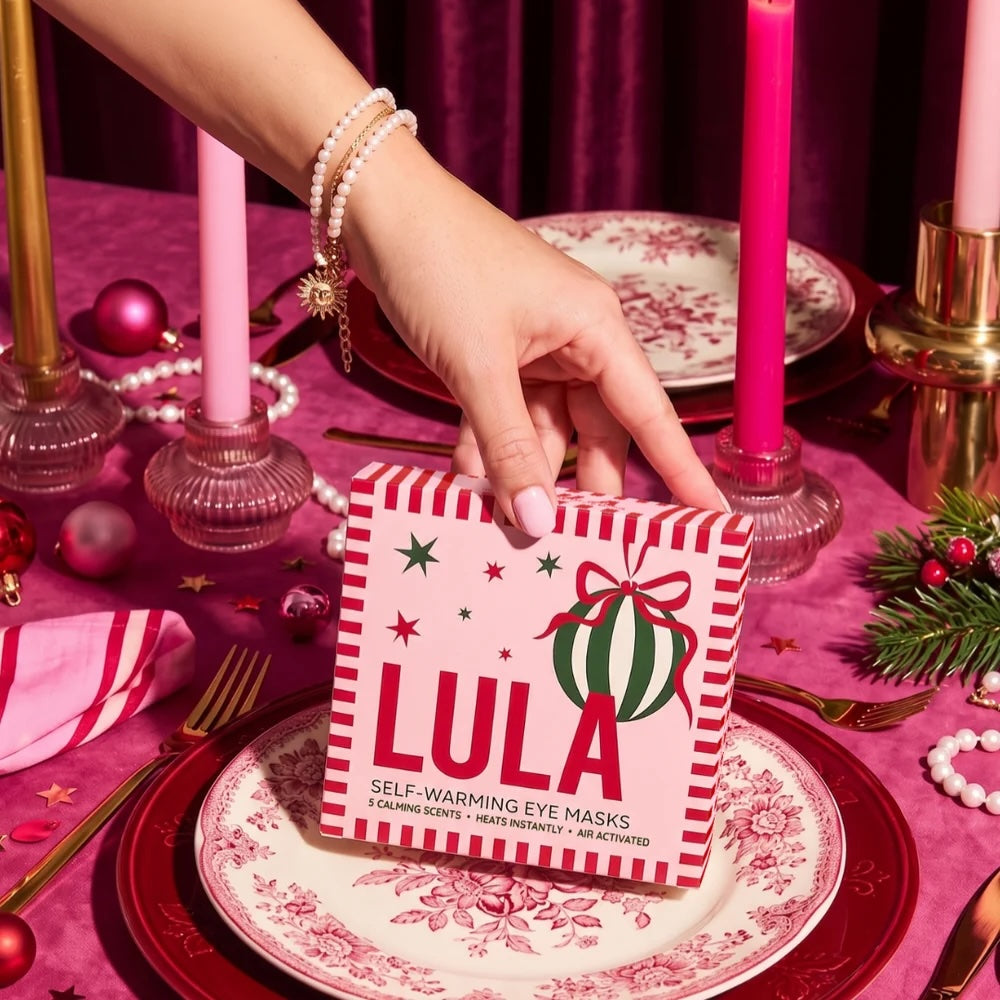 A hand holding a box of Self-Warming Eye Masks decorated with a Christmas theme, set on a decorated table with pink tablecloth, patterned plates, candles, and holiday greenery.