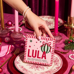A hand holding a box of Self-Warming Eye Masks decorated with a Christmas theme, set on a decorated table with pink tablecloth, patterned plates, candles, and holiday greenery.