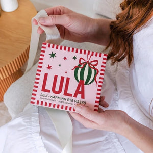 A person in a white robe holds a pink box of self-warming eye masks with a red-and-white striped border and a green ornament illustration. The scene is photographed in a bright indoor setting with soft natural light.