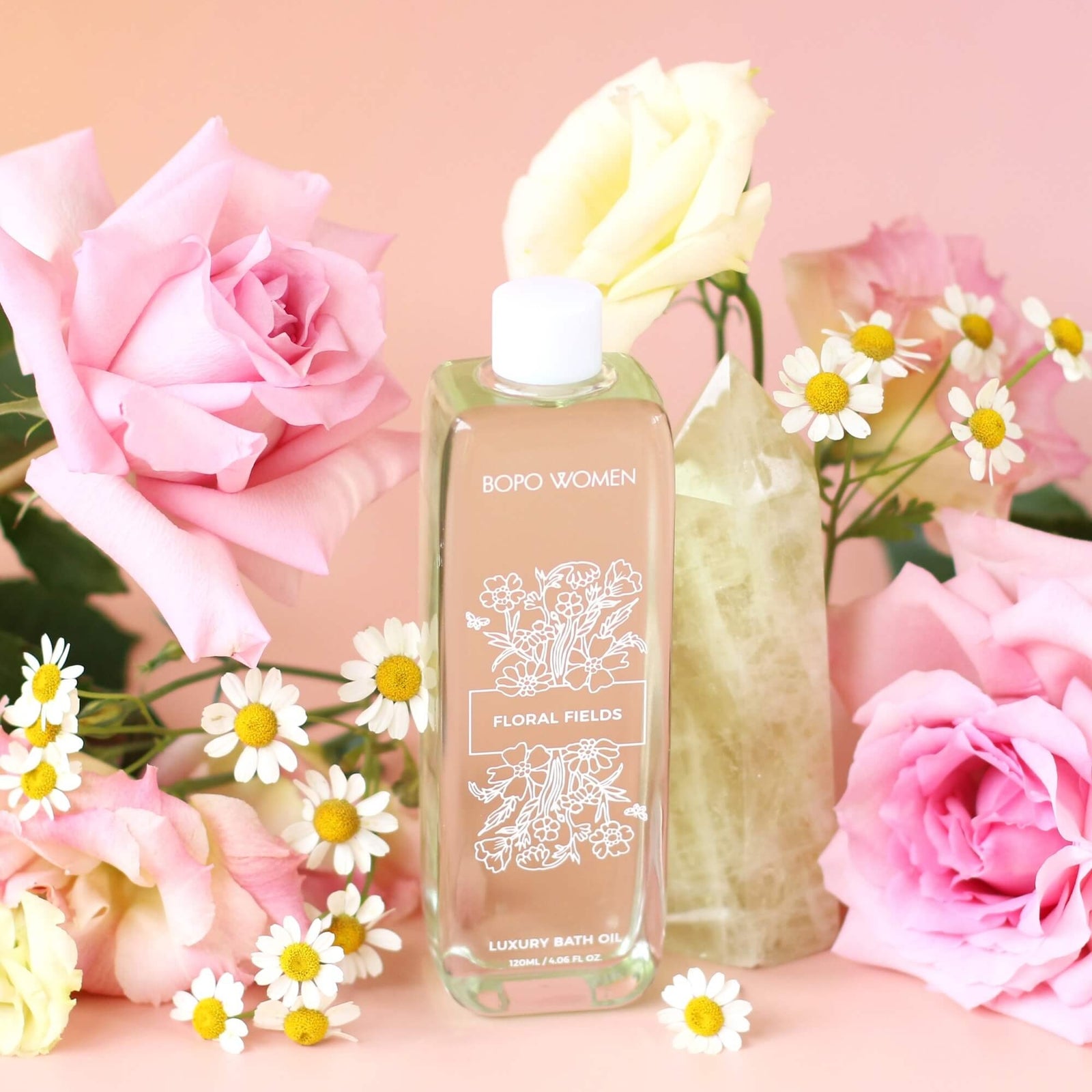 Bottle of floral fields luxury bath oil surrounded by pink roses and daisies on a pink background
