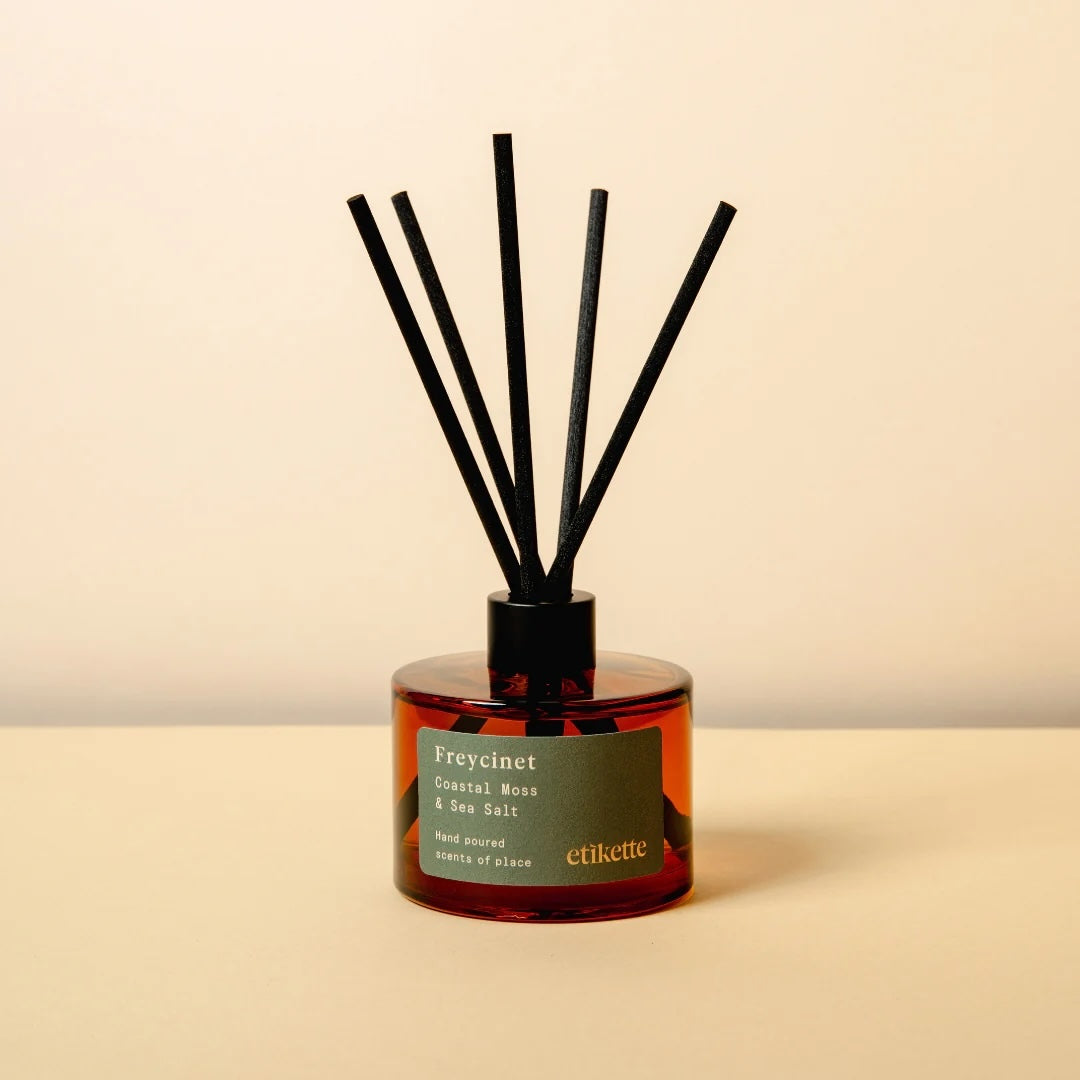 An amber glass jar candle with two lit wicks and a cream label reading 'Freycinet Coastal Moss & Sea Salt', set on a beige surface against a matching beige background.