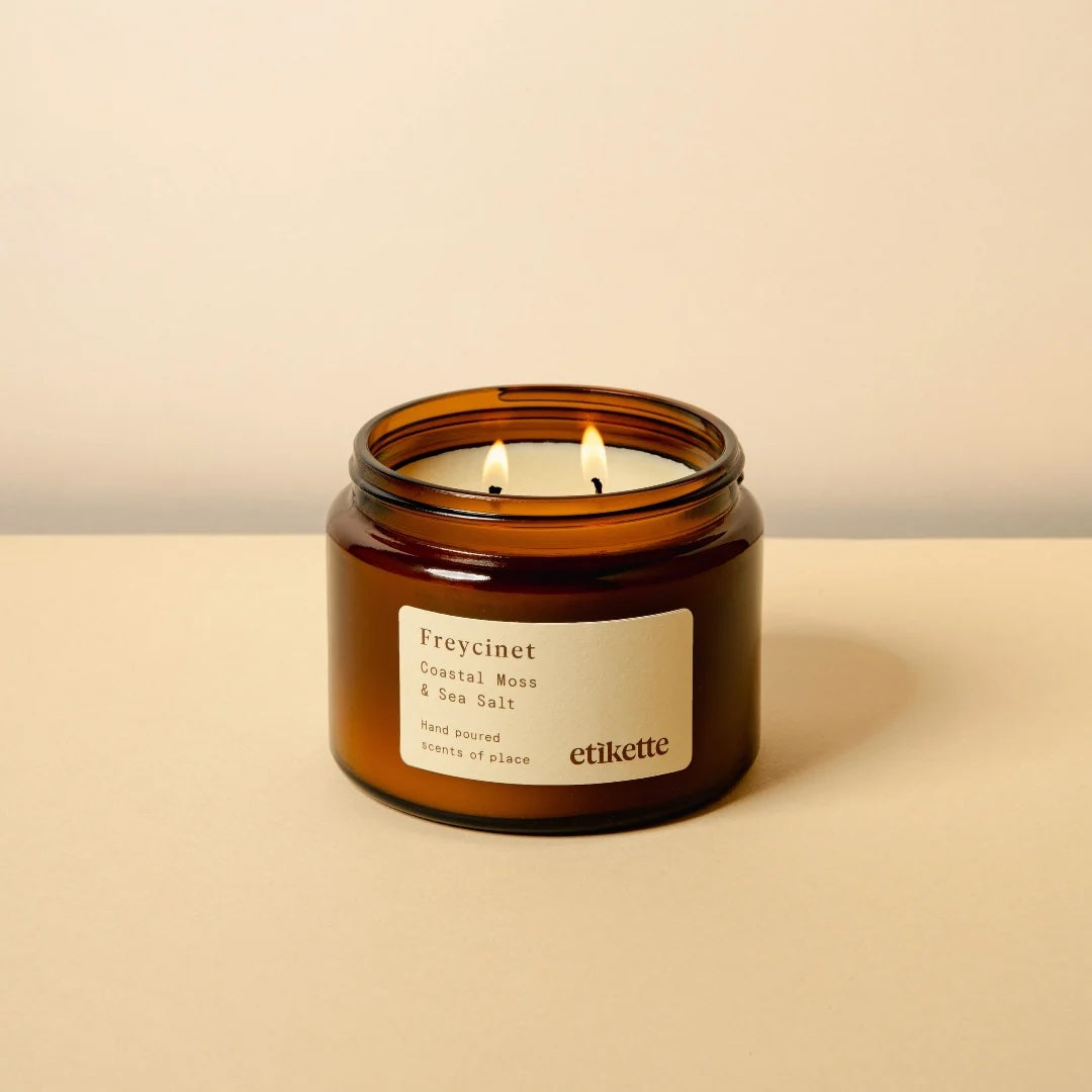 An amber glass jar candle with two lit wicks and a cream label reading 'Freycinet Coastal Moss & Sea Salt', set on a beige surface against a matching beige background.