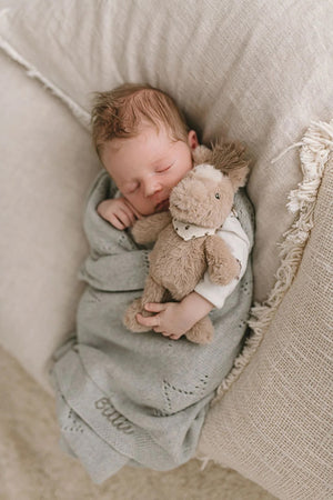 Sleeping baby holding a soft horse toy rattle in honey coloured plush fabric with a cream patterned neck bandana, on a neutral background
