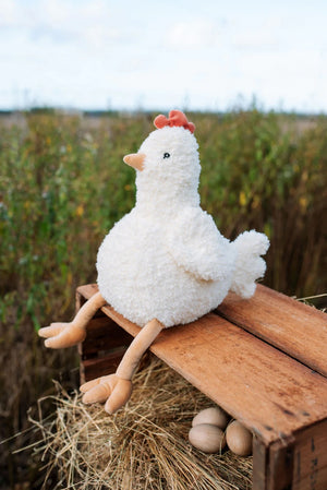 Plush soft toy chicken on a wooden coop with three eggs and a field in the background
