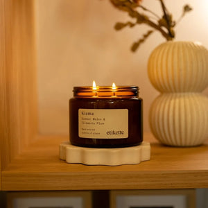 An amber glass jar candle with two lit wicks sits on a scalloped cream ceramic base on a wooden shelf. The label reads 'Klama' and 'etikette'.