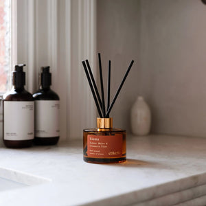 A reed diffuser bottle with amber liquid, a gold collar, and black reeds sits on a white marble countertop in a bathroom setting, with two matching bottles blurred in the background.