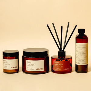 A four-piece set of Kiama scented candles and diffuser on a warm beige background, including two amber glass jars with black lids, a reed diffuser bottle with black reeds, and a tall amber bottle with a black cap.