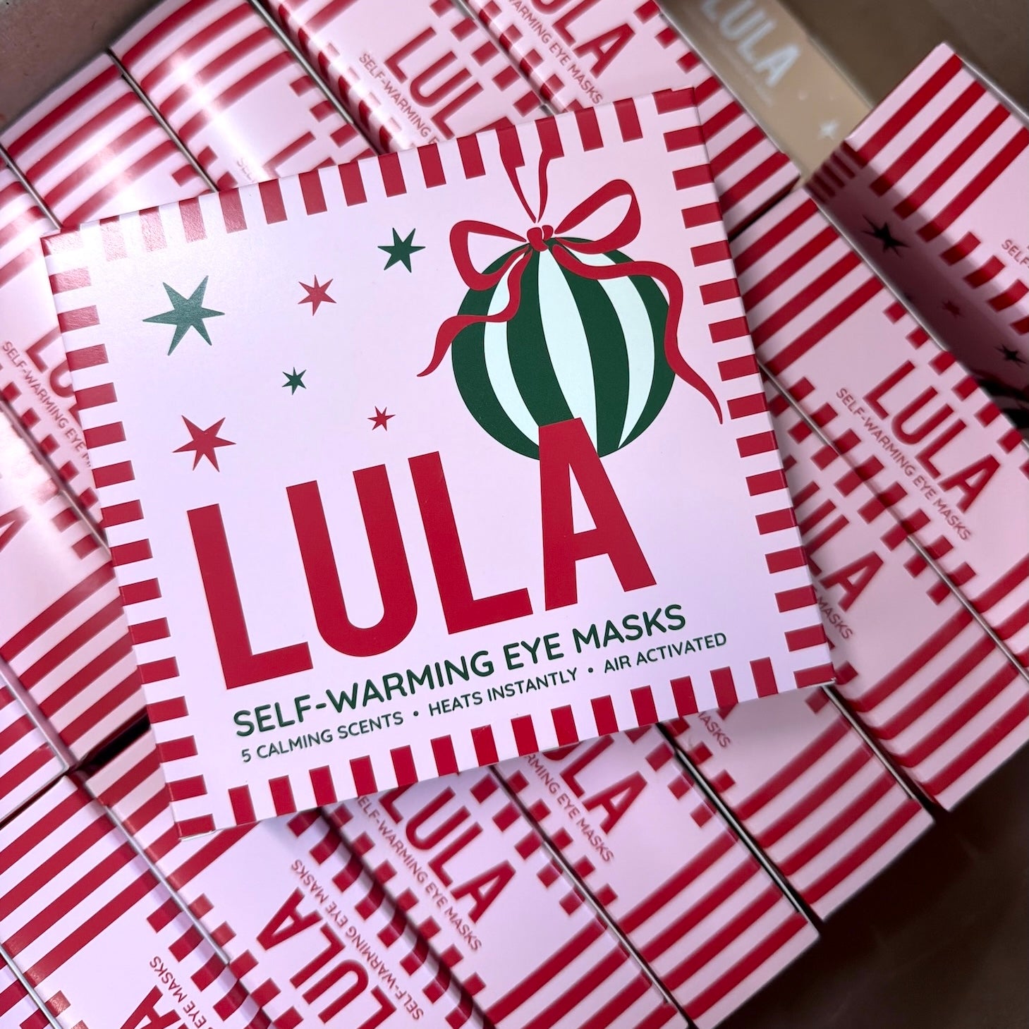 A hand holding a box of Self-Warming Eye Masks decorated with a Christmas theme, set on a decorated table with pink tablecloth, patterned plates, candles, and holiday greenery.
