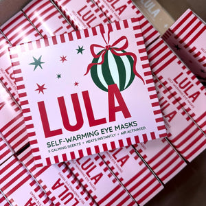 A stack of self-warming eye mask boxes arranged diagonally. The top box is white with red striped borders, a green and white striped ornament graphic with a red bow.
