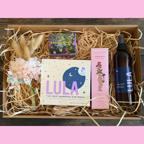 Gift pack of self-care products including warming eye mask, luxury hand cream, floral shower steamer, and magnesium spray bottle on a pink background