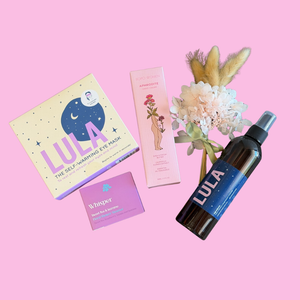 Gift pack of self-care products including warming eye mask, luxury hand cream, floral shower steamer, and magnesium spray bottle on a pink background